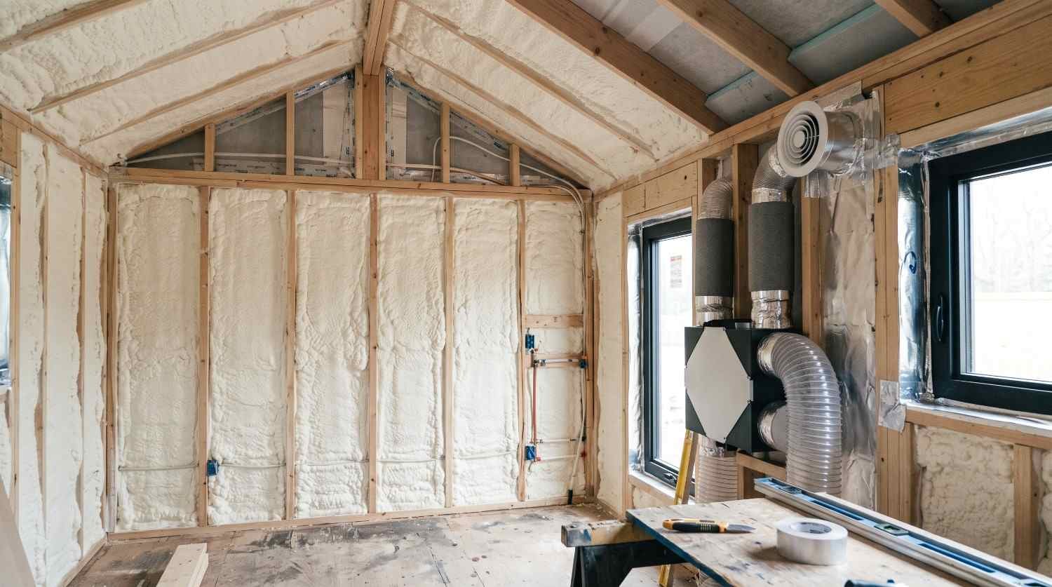 Spray foam insulation installed between the wooden wall and roof framing of a tiny home, with HVAC ventilation ducts and an air distribution system visible during construction.
