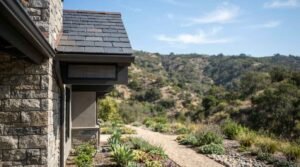 Exterior view of a custom home featuring fire-resistant stone veneer masonry, dark slate roof shingles, copper flashing, and ember-resistant mesh venting against a dry hillside landscape backdrop.