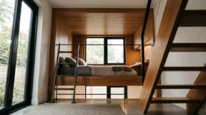 Modern ADU interior showcasing a built-in wooden loft bed nook with an attached metal ladder, integrated workspace, and floor-to-ceiling windows overlooking a garden