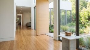Modern sustainable entryway featuring warm bamboo hardwood flooring, an open wood pivot front door, a marble accent pedestal table with small decor objects, and large floor-to-ceiling windows overlooking a green outdoor garden.