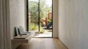 A floating light oak shelf displaying marble, wood, and brass finish samples in a minimalist hallway opening onto a private patio garden through a black steel glass door