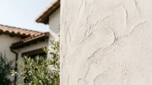 Close-up view of a textured white stucco exterior wall on a Mediterranean-style home featuring traditional roof tiles and green foliage.