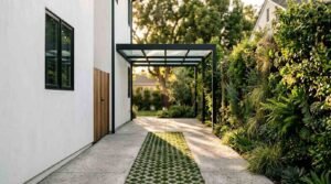 A modern concrete driveway with a central permeable grass grid runner leading to a black metal cantilevered glass carport, flanked by a white stucco home and a dense vertical living green wall