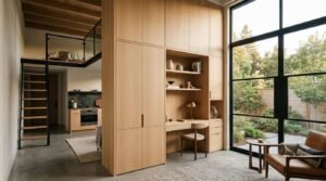 Modern ADU interior featuring custom floor-to-ceiling light wood cabinetry with a built-in desk, a sleeping loft with black metal stairs, and large glass doors looking out to a garden patio