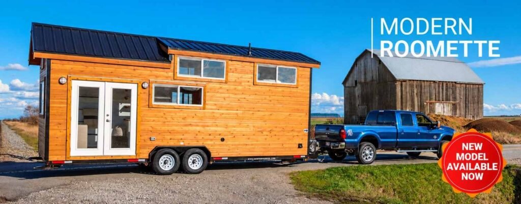Modern tiny home trailer with wood siding being towed by a blue pickup truck in a rural setting.