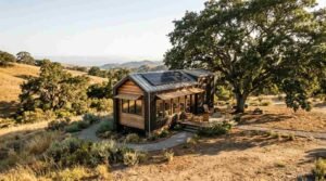 Solar-powered tiny home oriented on a scenic hillside with wooden deck and gravel path under large oak tree
