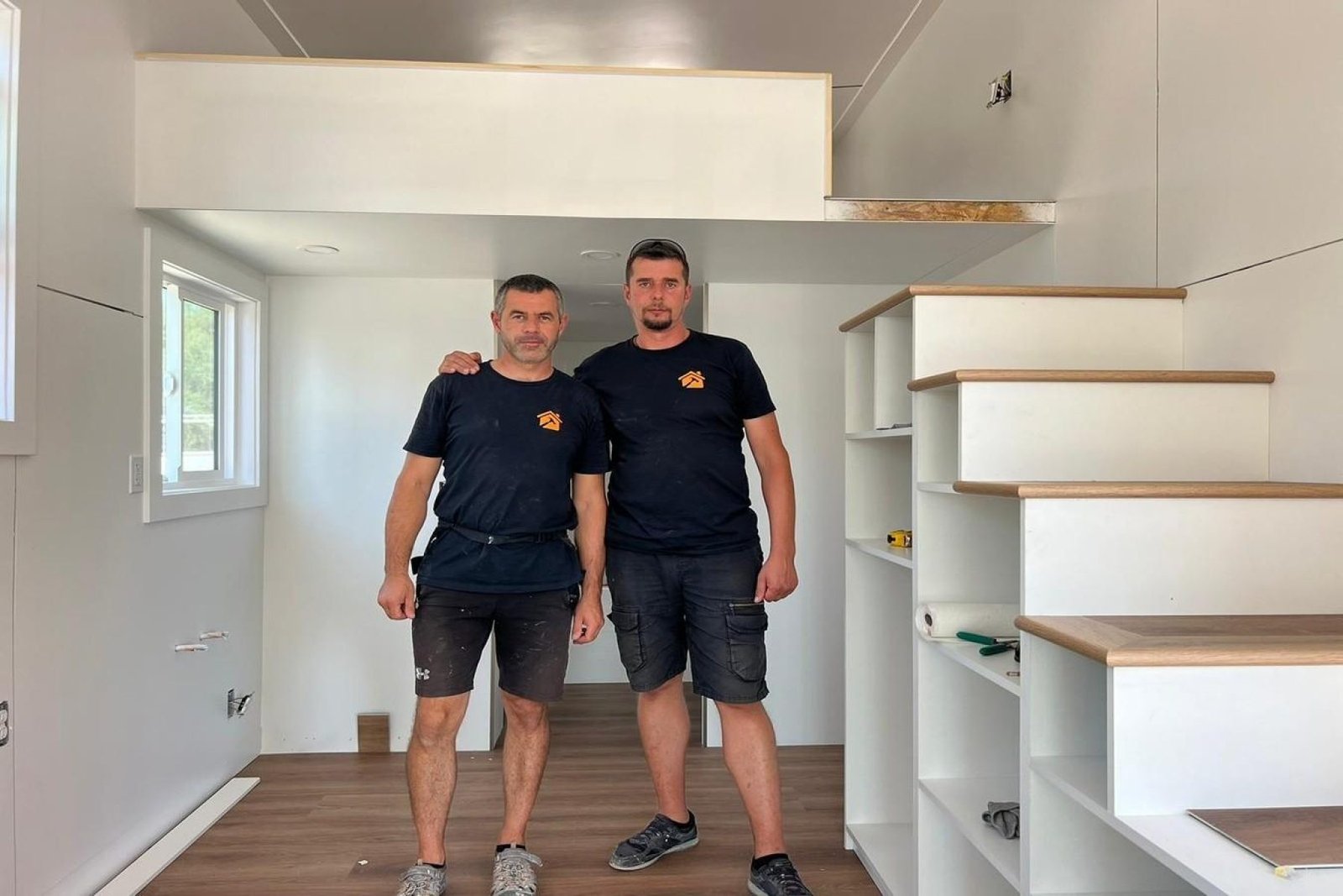 Tiny home builders standing inside a nearly finished tiny house with loft and custom staircase.