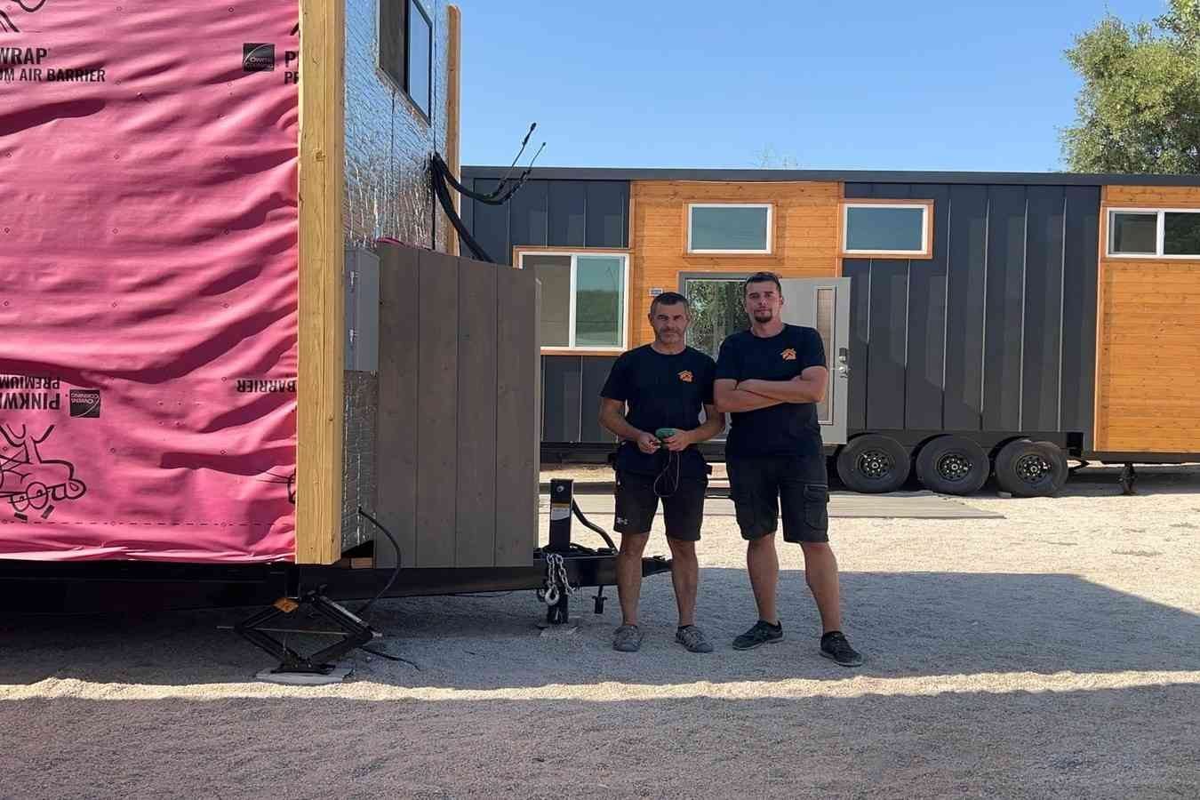 Tiny home builders standing beside a tiny house under construction at an outdoor fabrication yard.