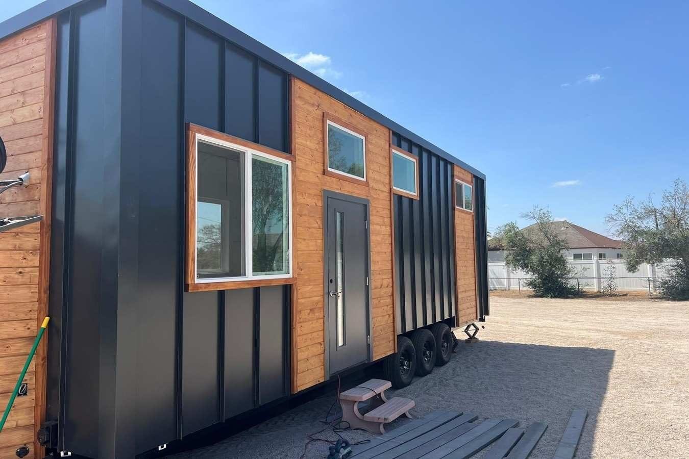 Modern tiny house with black metal siding and natural wood accents on a triple-axle trailer in an outdoor lot.