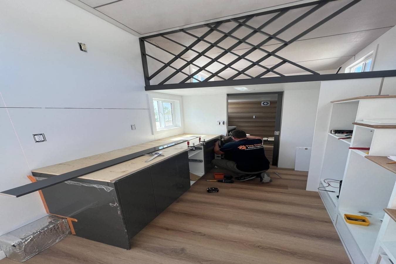 Builder installing kitchen cabinets inside a modern tiny home with loft, built-in shelving, and wood-look flooring.