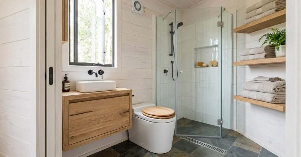 Tiny house bathroom with a glass-enclosed shower, floating wood vanity, compact toilet, and space-saving open shelving.